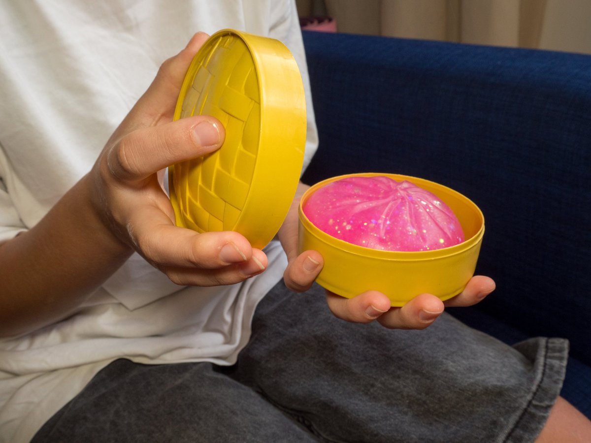 A child's hands unboxing a pink glittery squishy dumpling toy from a yellow steamer basket